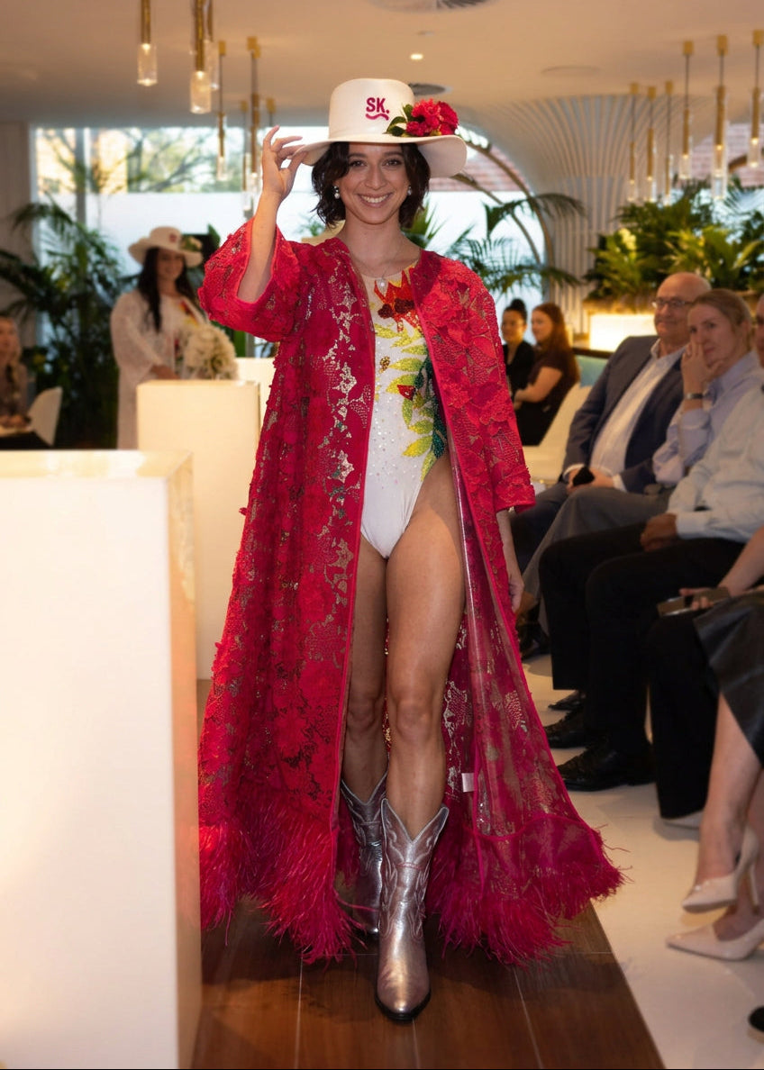 A woman walks down a fashion runway wearing a bright pink feathered coat over a white and green patterned dress, with a white hat decorated with pink flowers. Guests are seated in the audience along the right side of the runway.