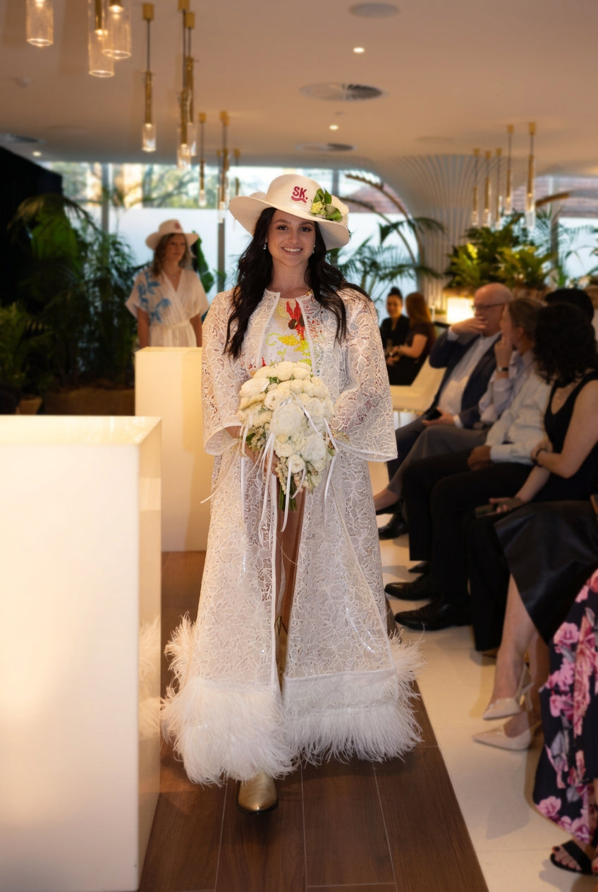 A woman walks down a runway wearing a long white lace and feather coat, a white cowboy hat with red lettering, and holding a bouquet of white flowers. An audience is seated along the side of the runway in a modern indoor venue with hanging lights and plants.