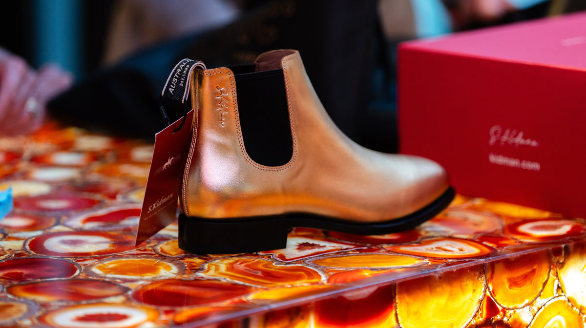 A metallic rose gold Chelsea boot with black elastic side panels and a black sole, displayed on a colorful circular-patterned surface next to a S. Kidman shoebox.