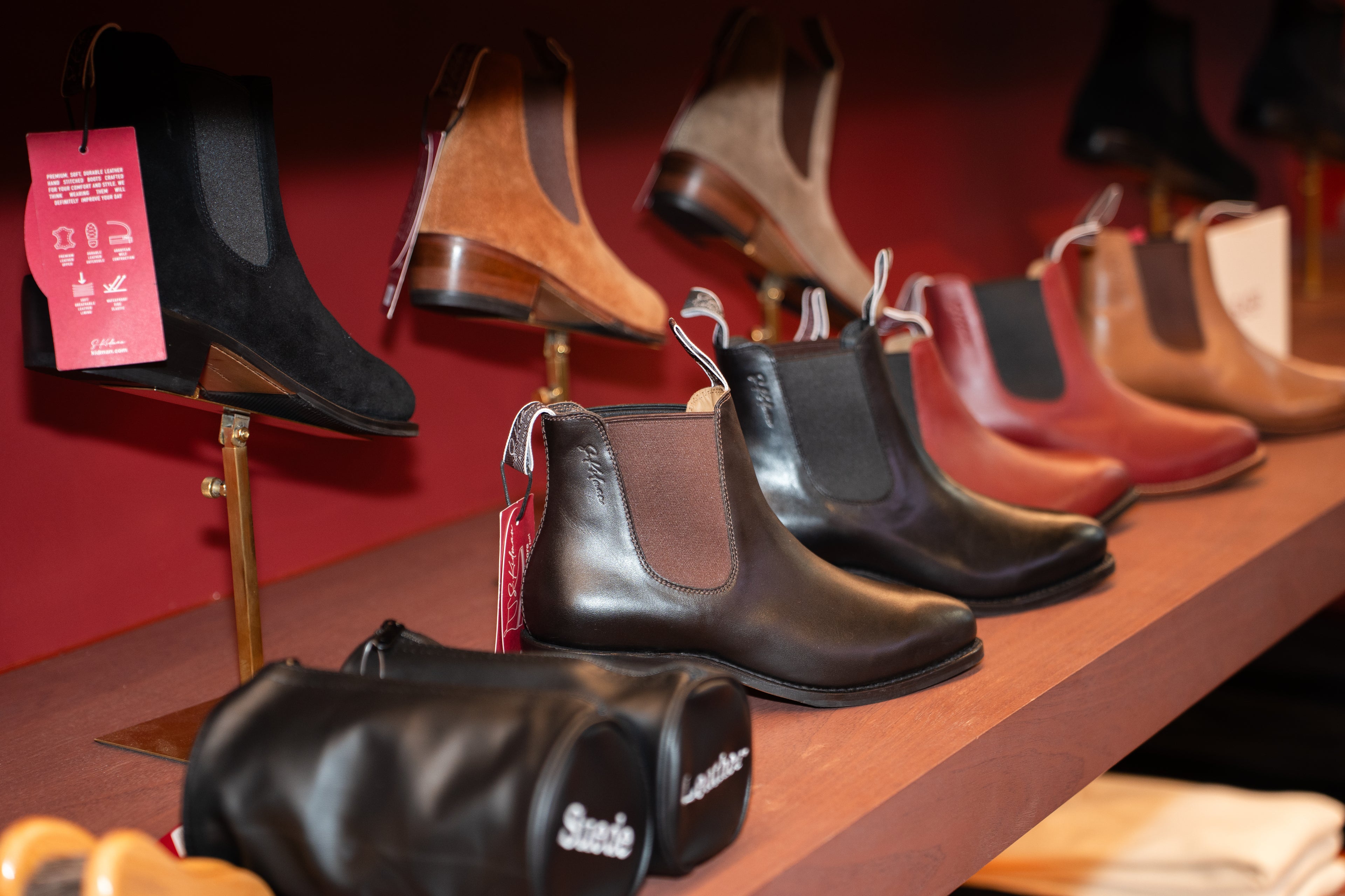 A display of Suede Chelsea boots in various colors including black, brown, and red, shown on wooden shelves against a dark red background.