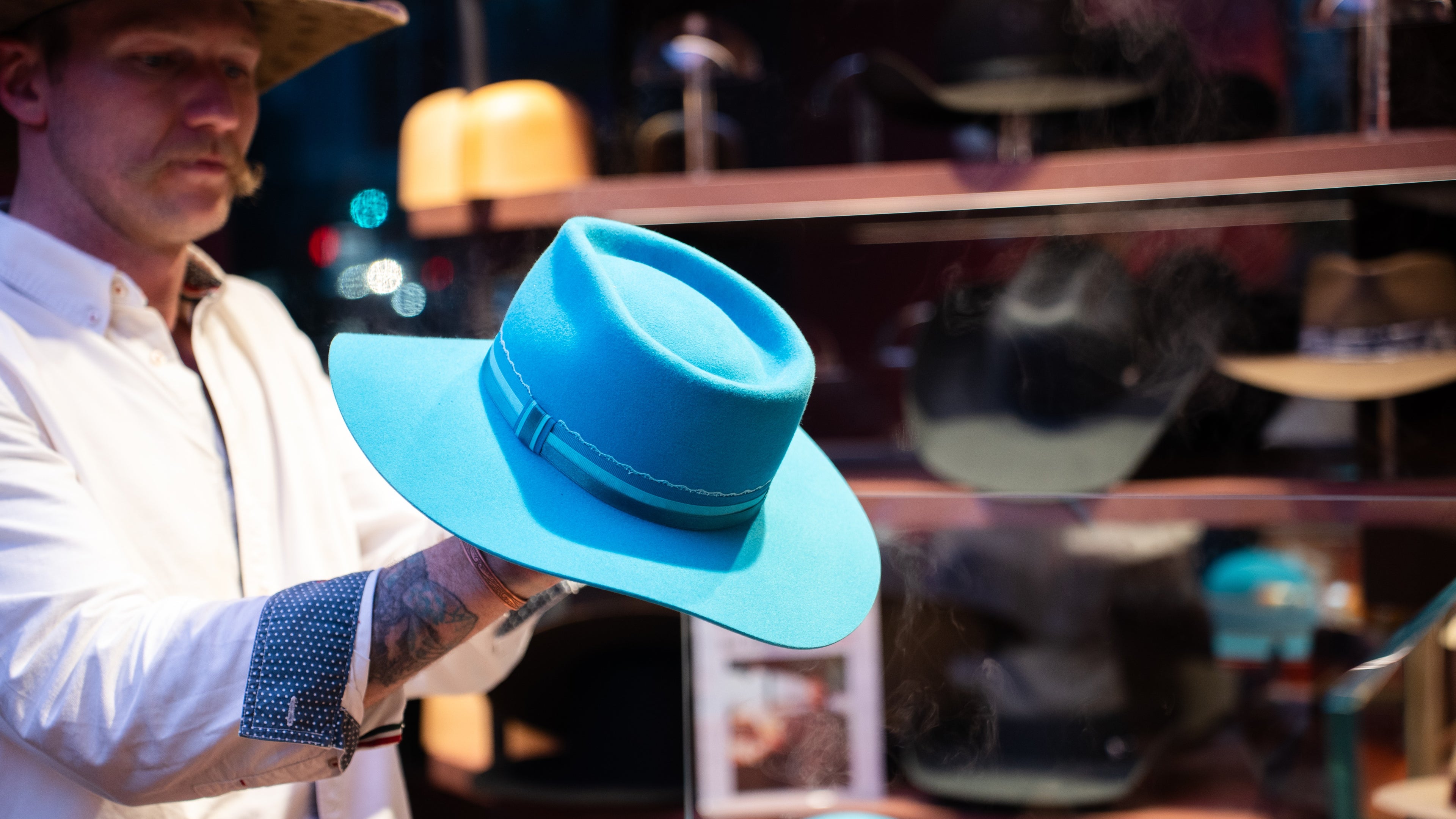 A man in a white shirt and straw hat holds up a bright turquoise wide-brim fedora hat in front of a blurred shop display.