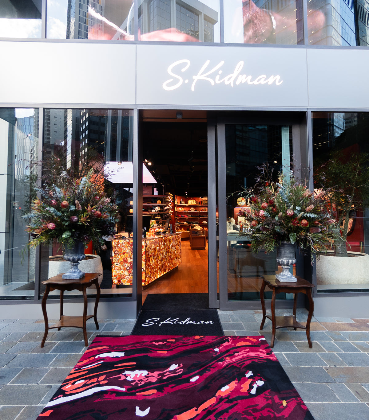 S.Kidman boutique storefront with glass doors, a white sign above, floral arrangements on both sides, a black branded doormat, and a bold red, black, and pink patterned runner rug on the pavement.