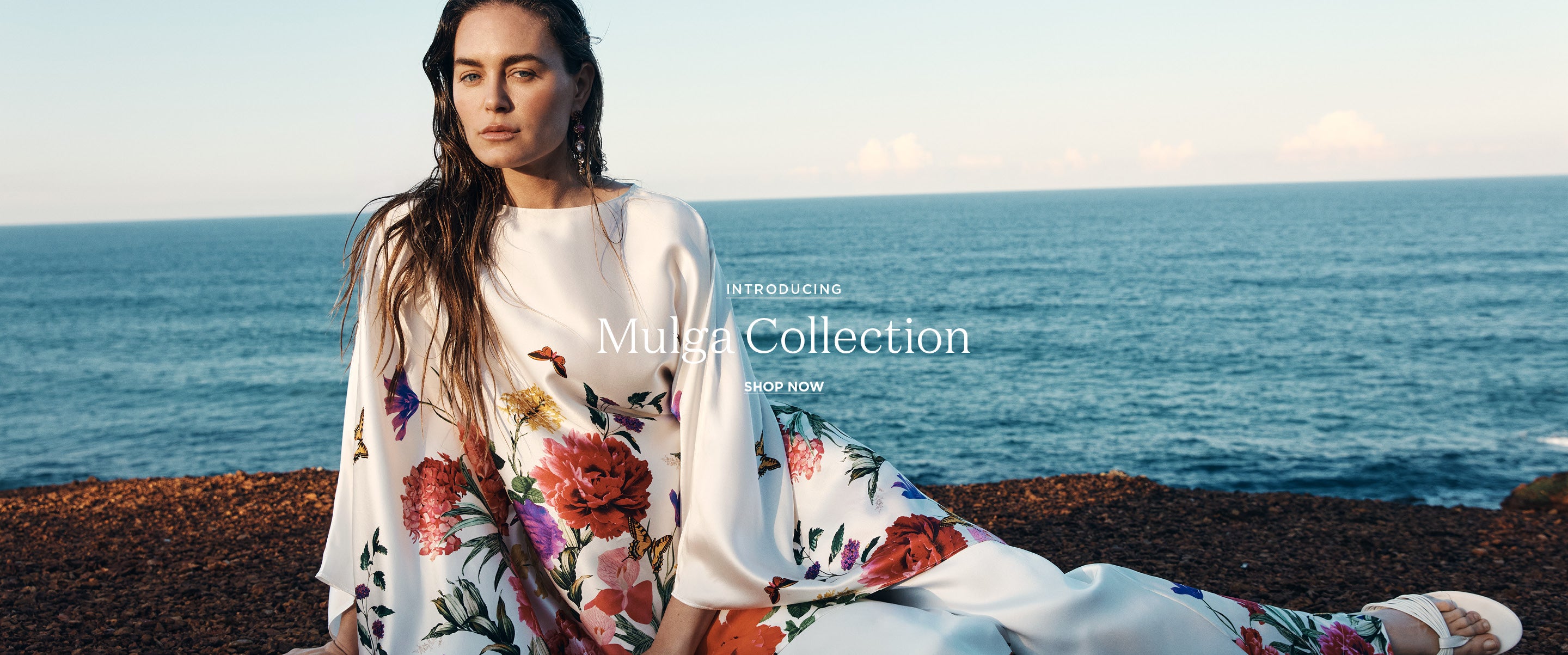 A woman sits on a rocky shoreline with the ocean and sky behind her, wearing a flowing white dress with large multicolored floral prints. Text on the image reads 'Introducing Mulqa Collection'.