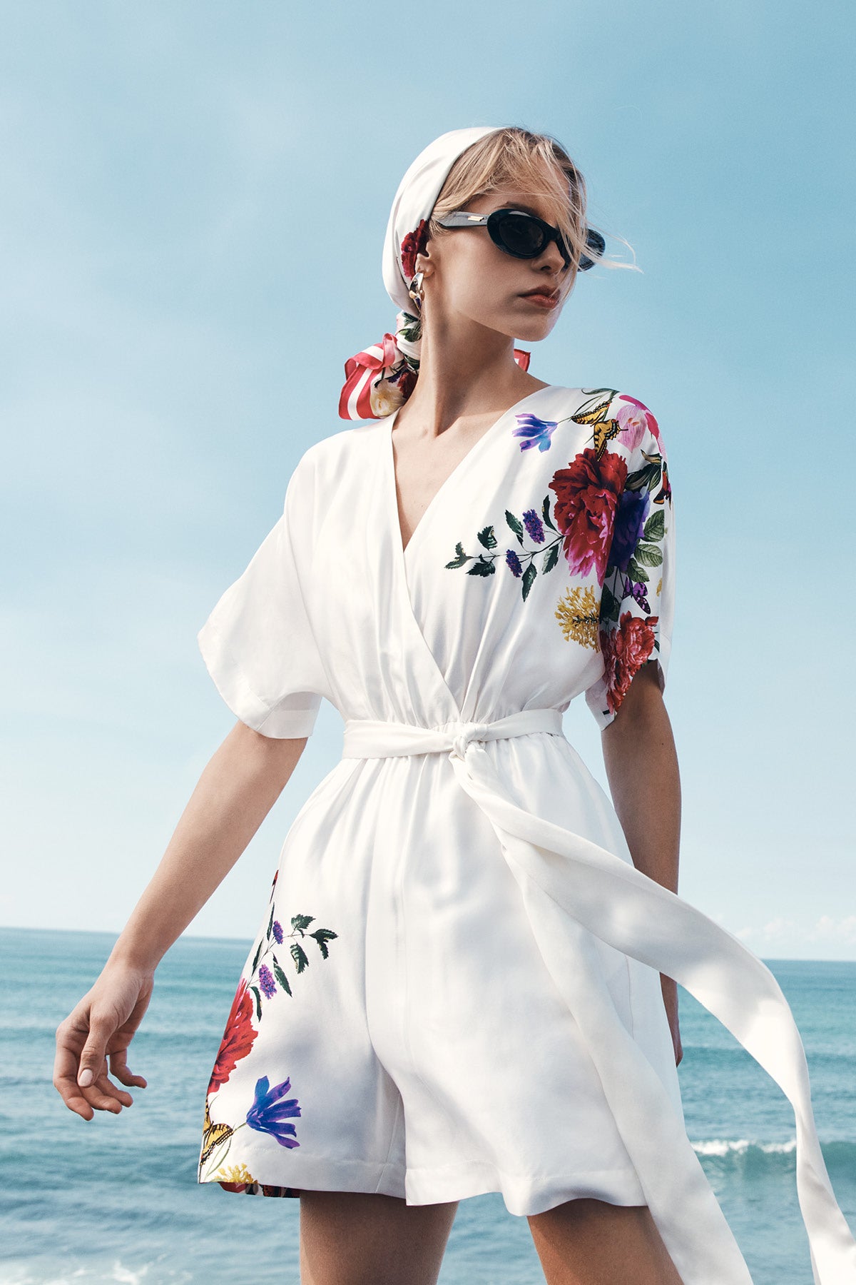 Mulga Collection by Kidman. A woman wearing a white belted wrap-style romper with colorful floral embroidery stands outdoors by the ocean under a blue sky. She is also wearing a white headscarf and dark sunglasses.