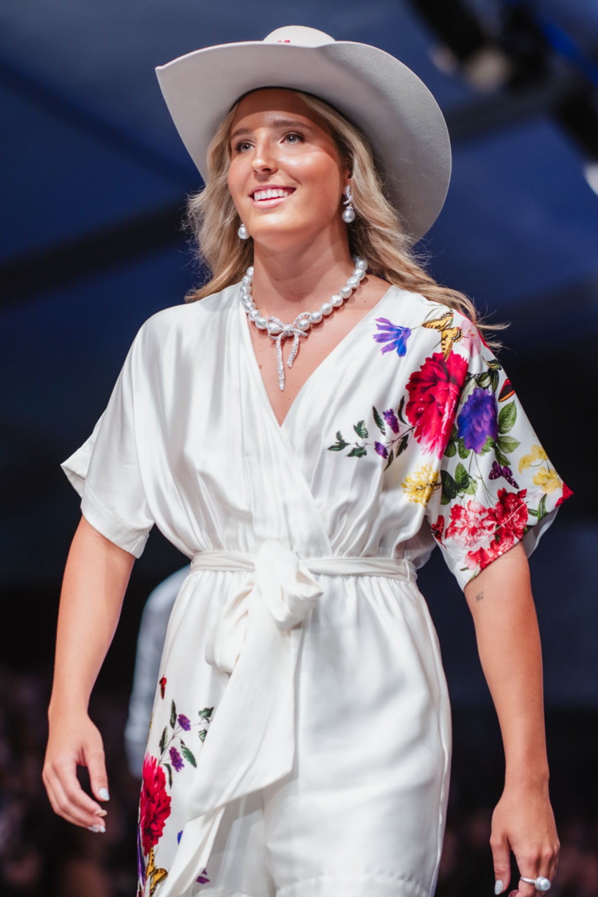 A woman wearing a white satin wrap-style romper with colorful floral embroidery on the sleeves and hem, a matching white wide-brim hat, and a pearl necklace, photographed on a runway or stage.