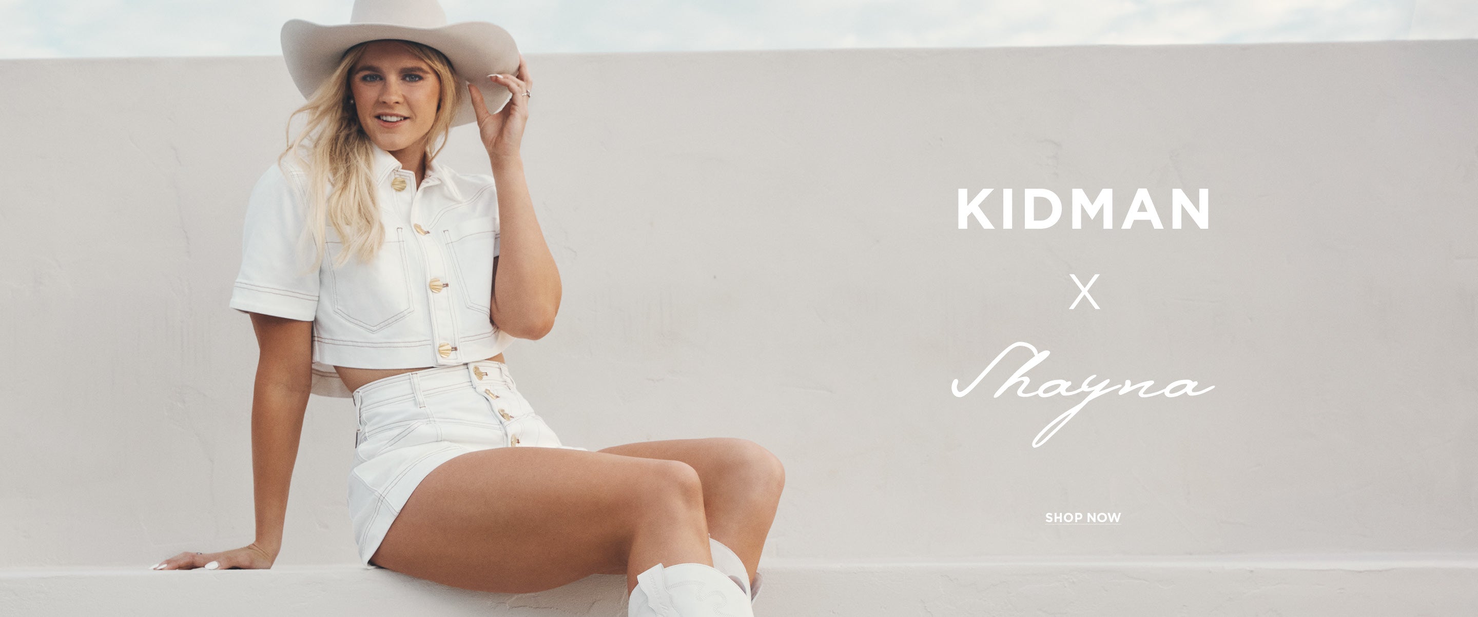 A woman wearing a white short-sleeve button-front shirt and matching white shorts sits against a light neutral wall, holding a cowboy hat. The right side features large white text reading 'KIDMAN x Shayna'.